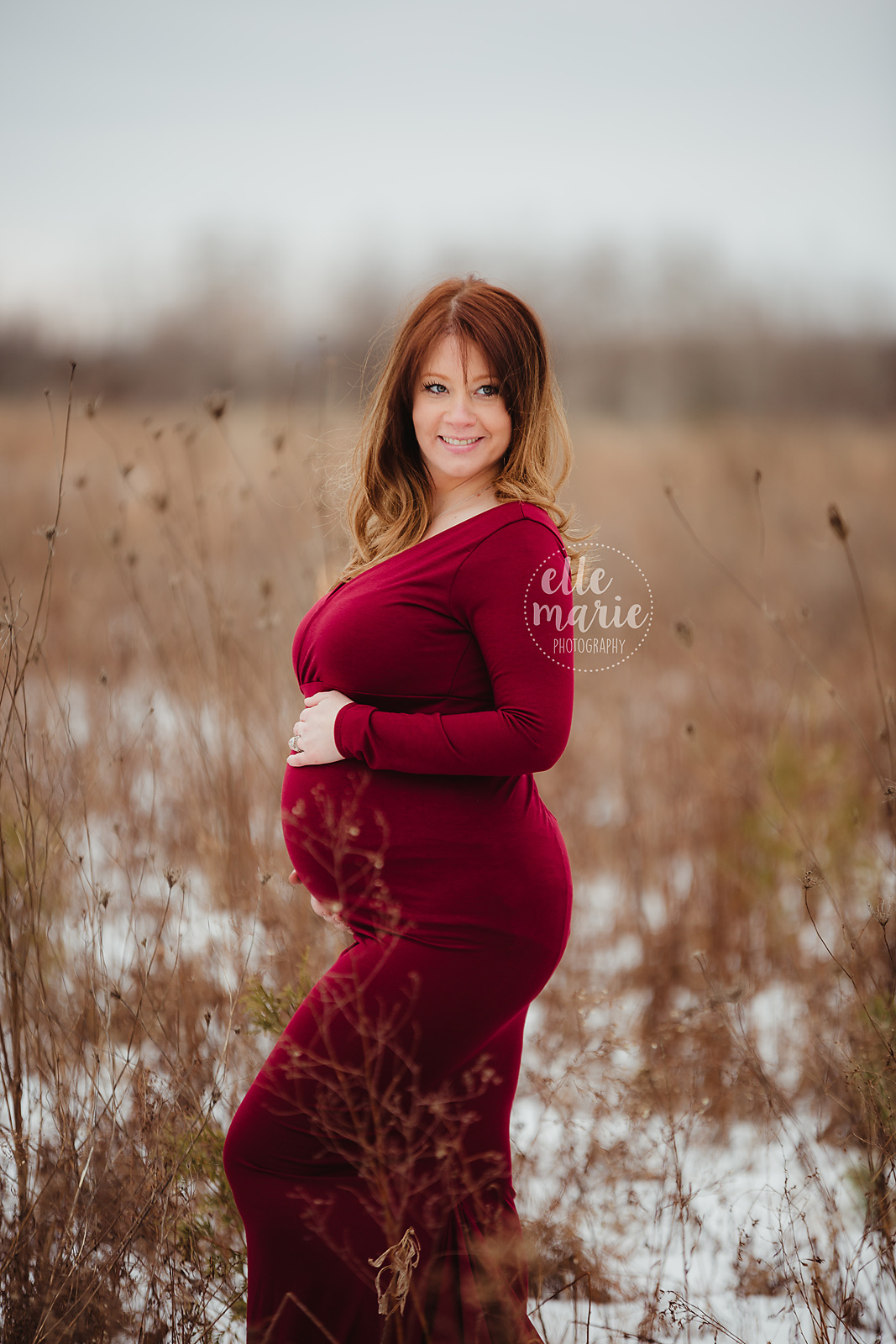 expecting mother in red gown in winter setting, smiling at husband off camera