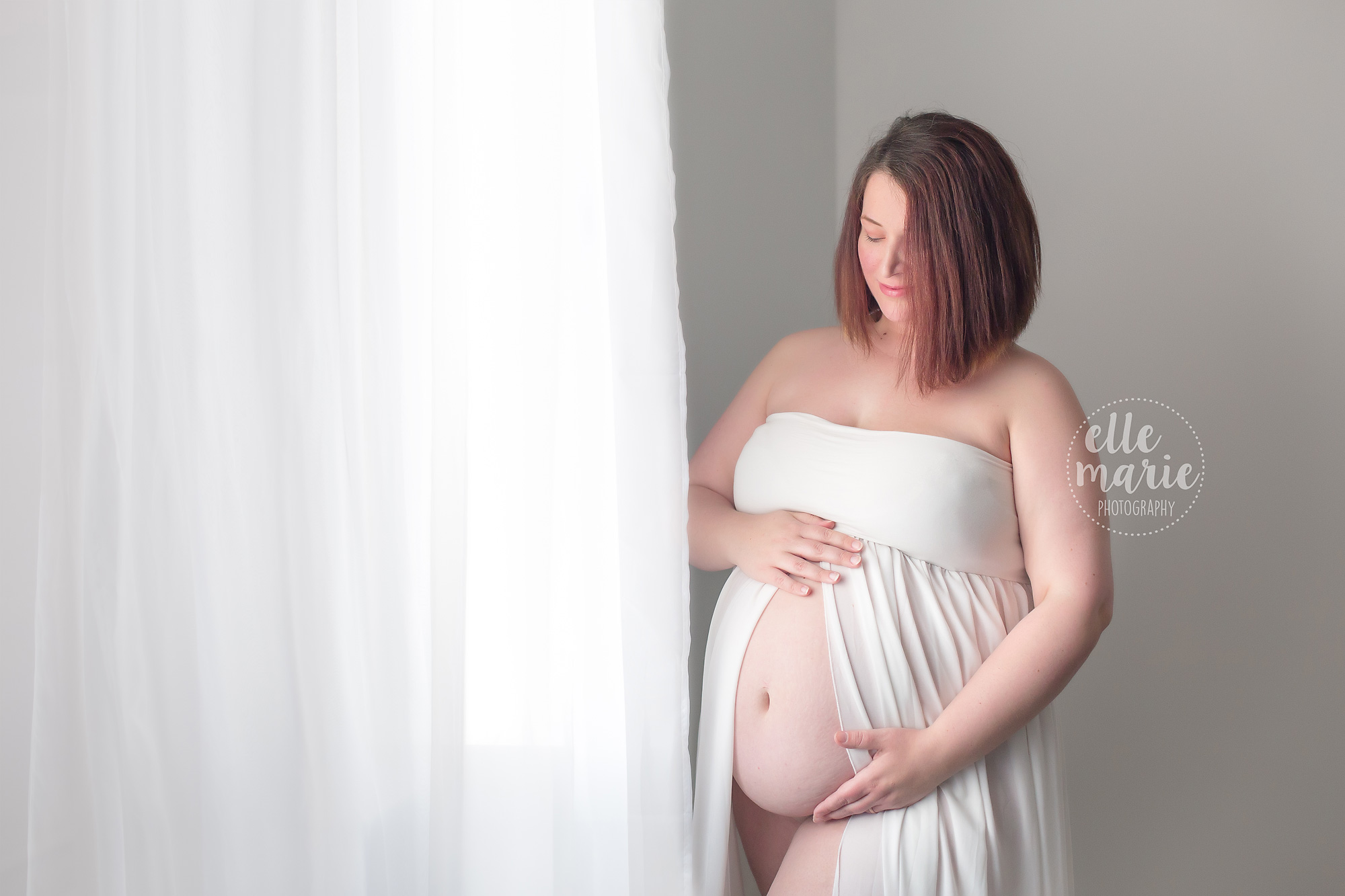 pregnant mother standing by window in white gown for maternity photos