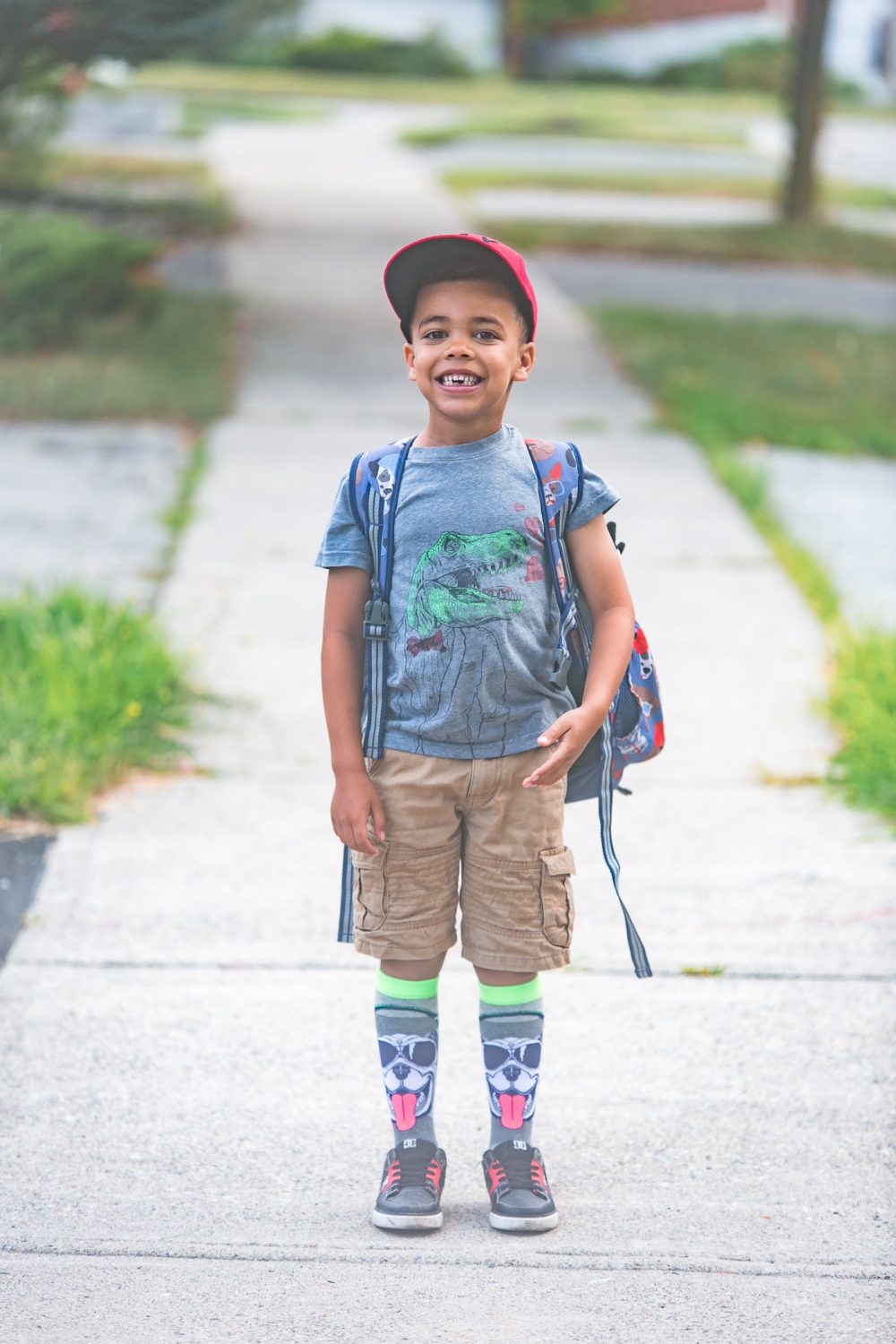 Young boy ready for first day of school, Wearing a red hat, grey shirt with a dinosaur and knee socks with a dog on them.