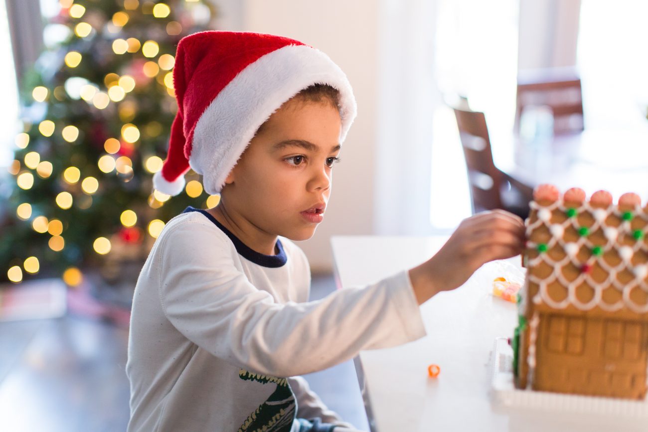 Young boy making a gingerbread house while wearing a santa hat