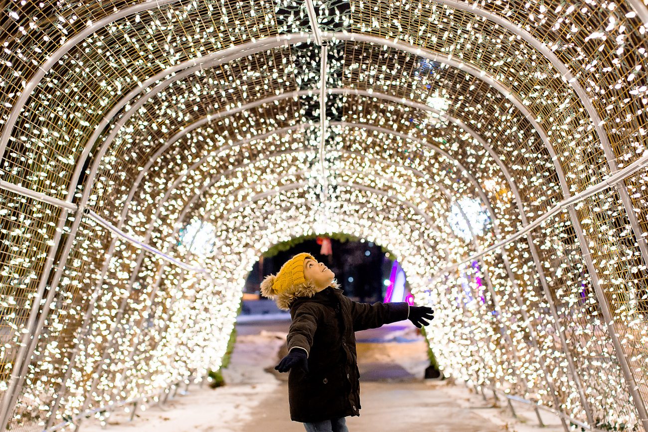 young boy stands under Christmas light archway wearing a yellow hat and spinning
