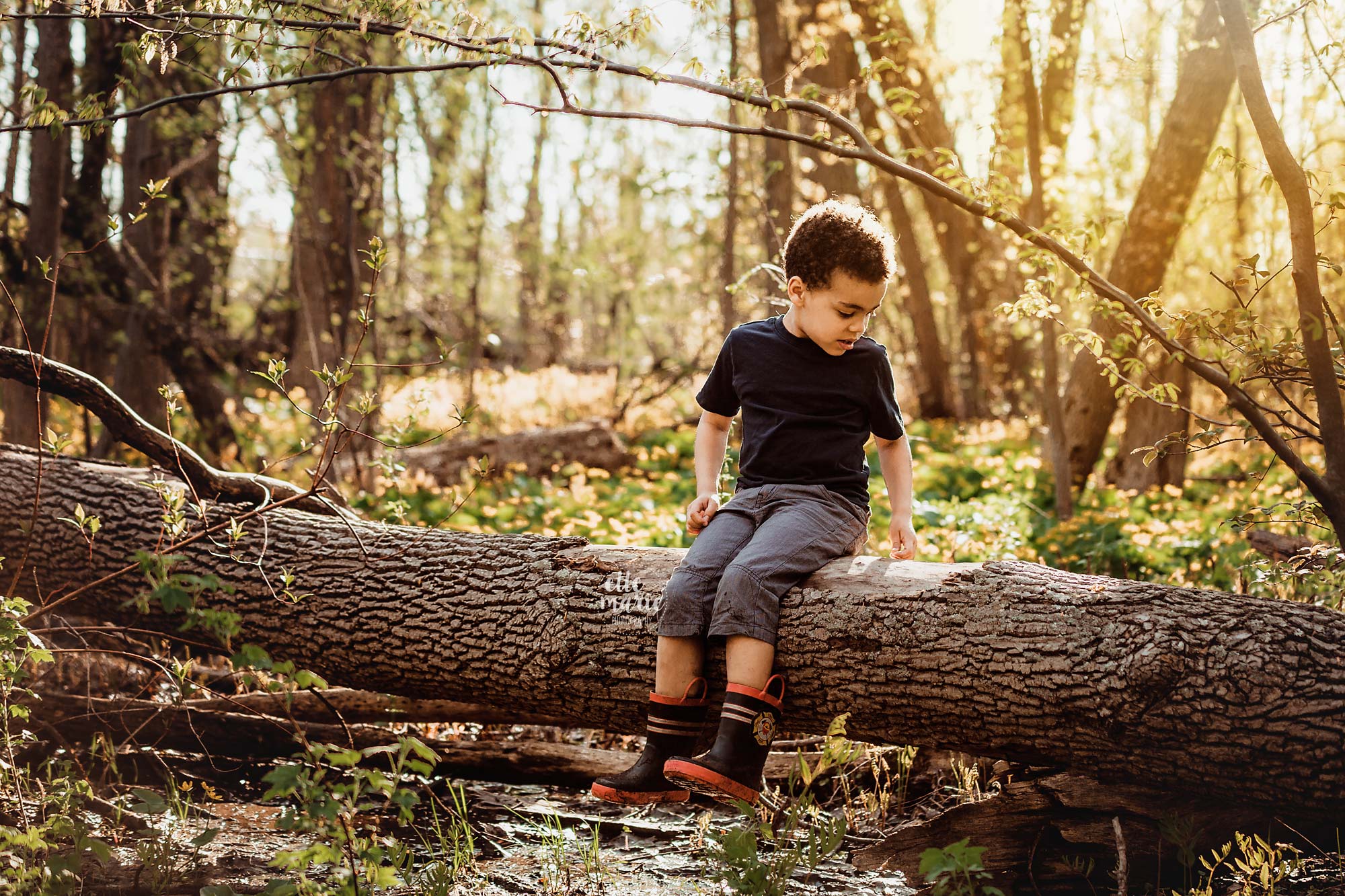 young boy wearing rubber boots sits on a log in the forest