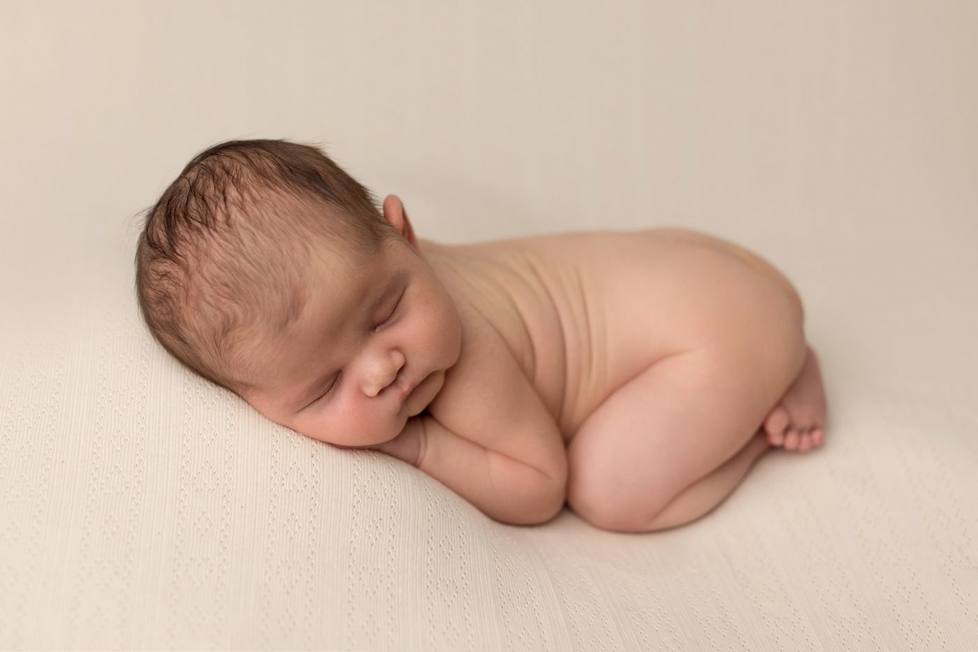 newborn baby curled up, laying on cream blanket