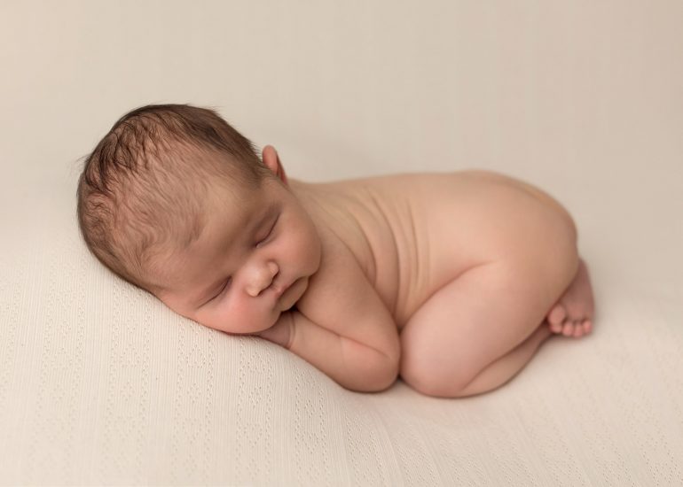 newborn baby curled up, laying on cream blanket