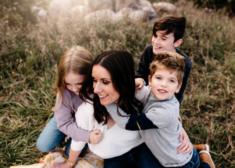 mom sitting in grass being hugged by her 3 children