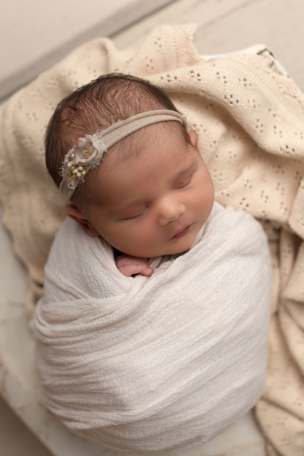 A newborn baby sleeps in a white swaddle on a cream blanket with a headband