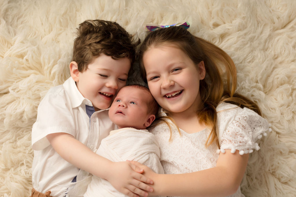 Two happy siblings laugh while laying on a fur blanket hugging their newborn baby sibling after using a 3d ultrasound whitby