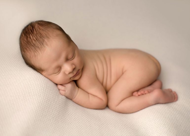 A newborn baby sleeps in froggy pose on a bean bag in a studio