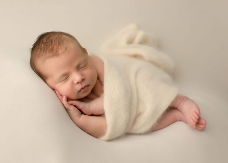 A newborn baby sleeps on its side on a white pad wrapped in a white blanket