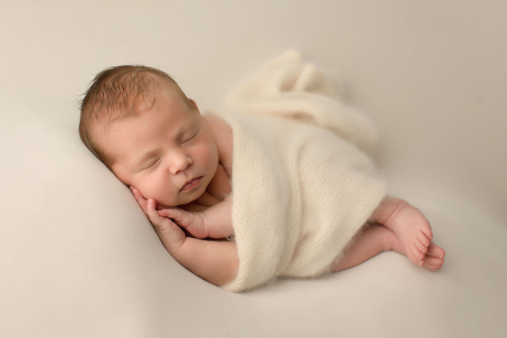 A newborn baby sleeps on its side on a white pad wrapped in a white blanket