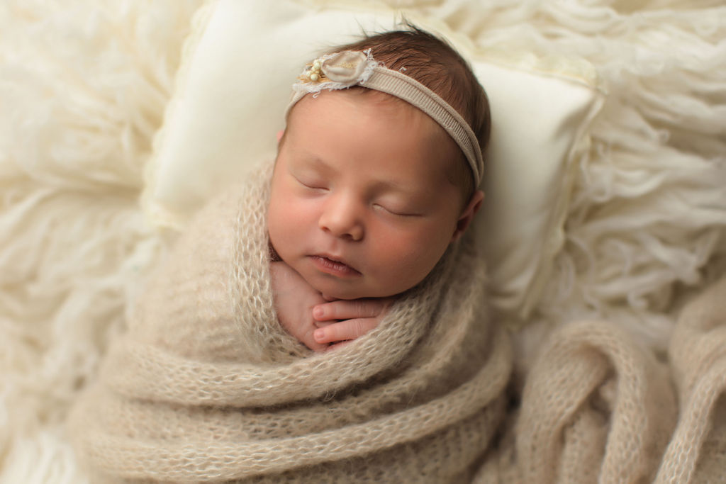 A newborn baby sleeps in a knit beige swaddle with a matching headband on a pillow and fur blanket