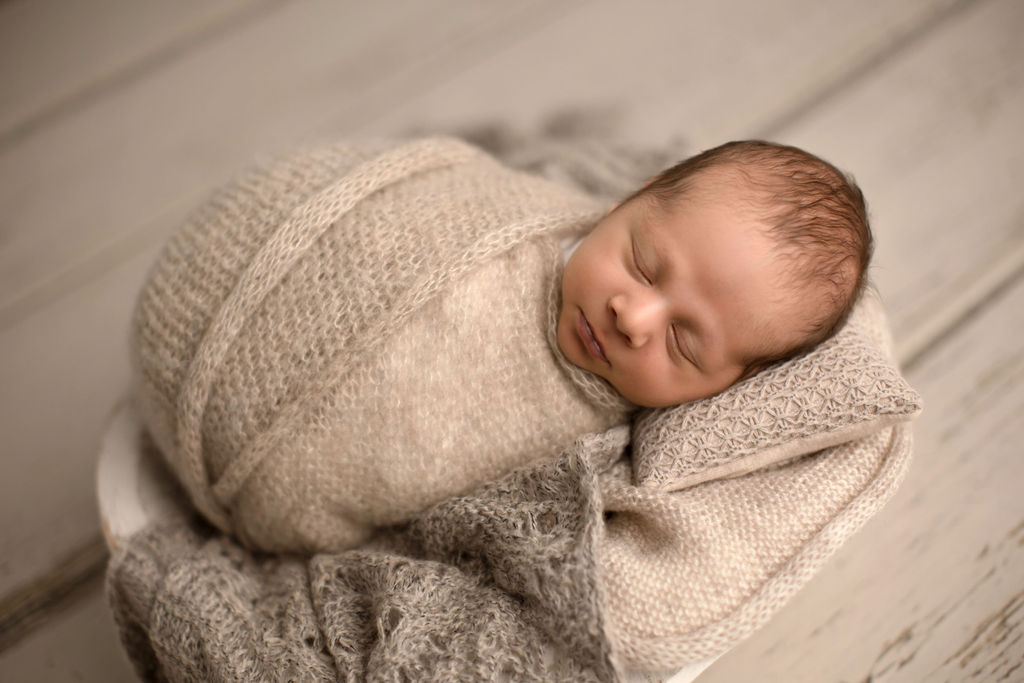 A newborn baby sleeps in a knit blanket in a wooden bucket on a wooden floor after visiting baby store oshawa