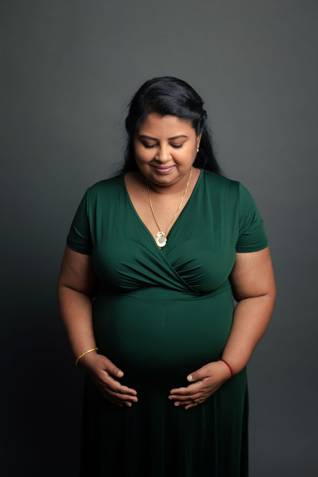 A mother to be in a green maternity gown smiles down to her bump while standing in a studio