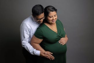 A father to be reaches to touch the bump while hugging his pregnant wife from behind in a studio after meeting community care midwives