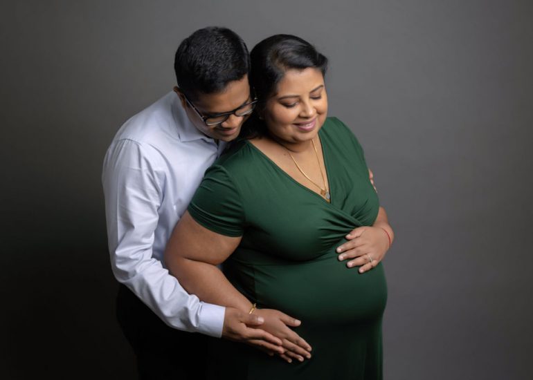 A father to be reaches to touch the bump while hugging his pregnant wife from behind in a studio after meeting community care midwives