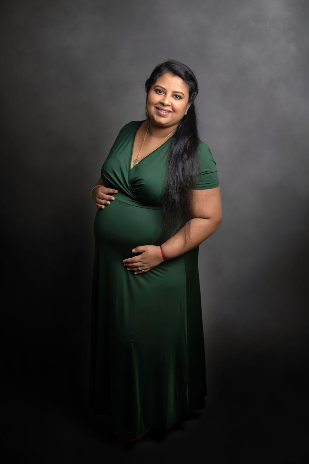 A mother to be in a green maternity gown stands in a studio holding her bump after meeting community care midwives