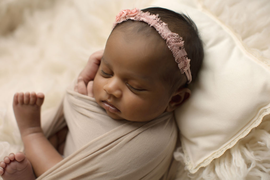A newborn baby girl in a pink headband sleeps on a pillow in a brown swaddle