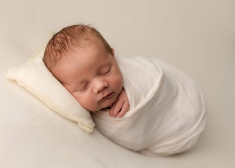 A newborn baby sleeps in a white swaddle with a hand poking out under its chin