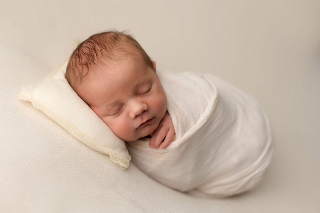 A newborn baby sleeps in a white swaddle with a hand poking out under its chin