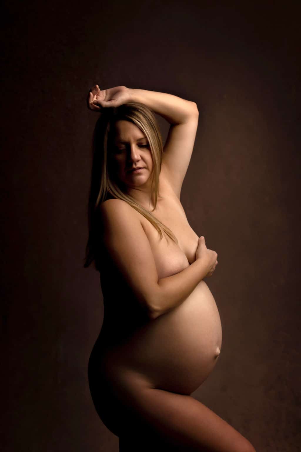 A mother to be stands in a studio with an arm above her head and the other covering her breast after meeting midwives oshawa