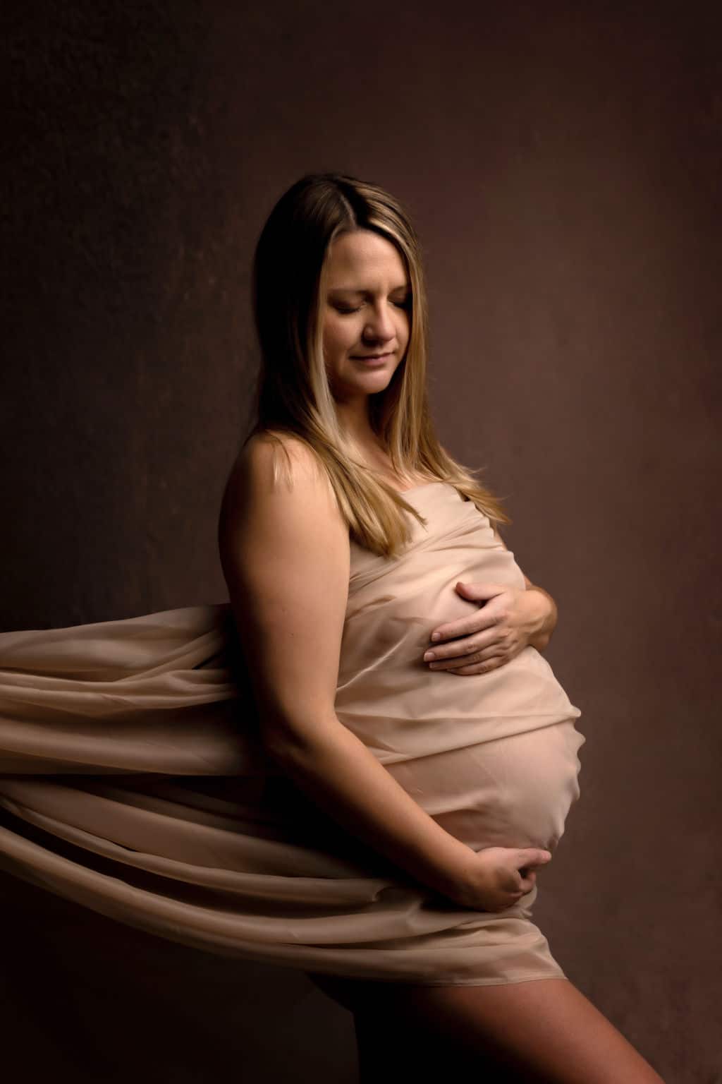 A mother to be wrapped in a beige fabric stands in a studio smiling down to her bump