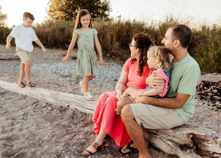 Two toddlers walk on a long piece of driftwood while mom and dad sit on it with their younger sister