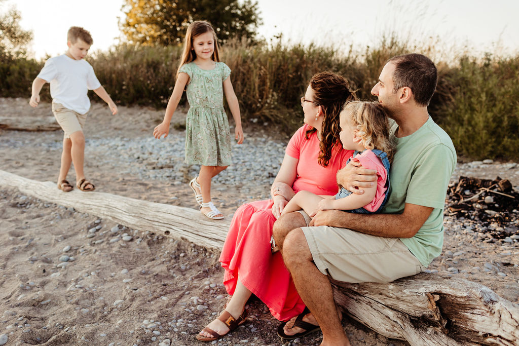 Two toddlers walk on a long piece of driftwood while mom and dad sit on it with their younger sister