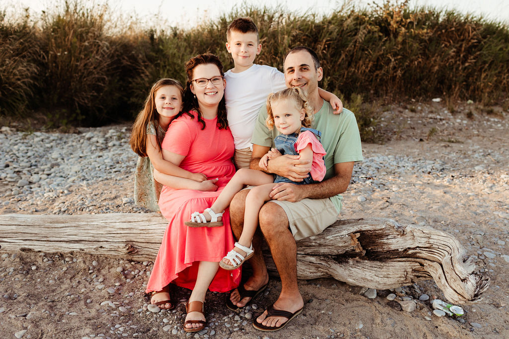 Happy parents sit on a piece of driftwood on a beach with their three toddler children hugging them after meeting obgyn whitby