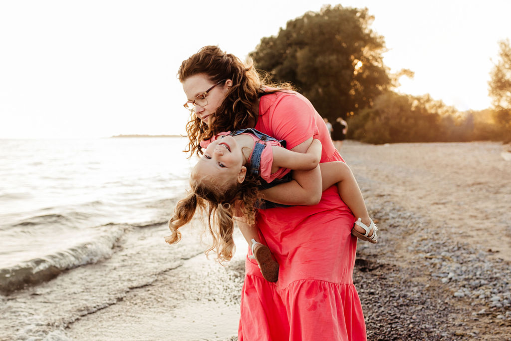 A mother in a pink dress tips her toddler daughter upside on a beach at sunet after using obgyn whitby