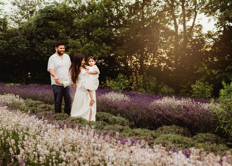 A young toddler girl in a white dress is carried by mom through a field of white and purple lavender at sunset with dad following behind