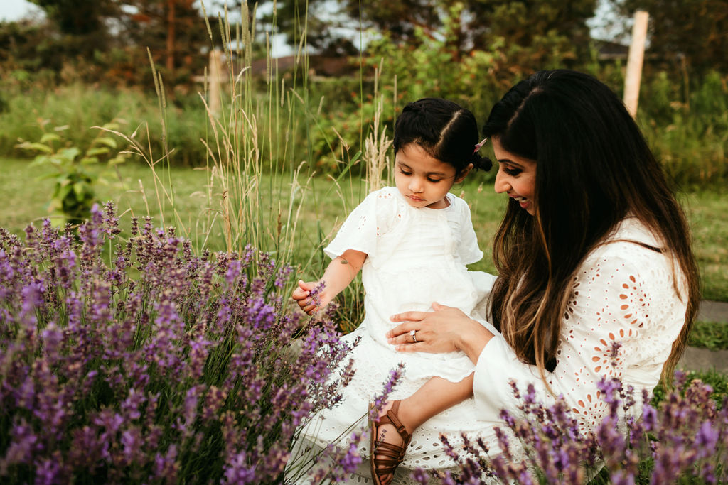 A mother in a white dress sits in a lavender garden exploring the flowers with her toddler daughter on her lap in a matching dress after visiting pediatrician whitby