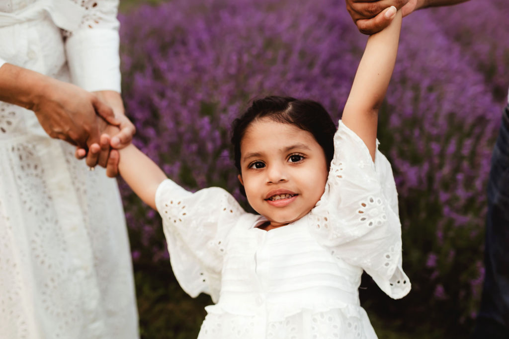 A young toddler girl in a white dress plays and swings with mom and dad holding her hands in a lavender field after visiting a pediatrician whitby