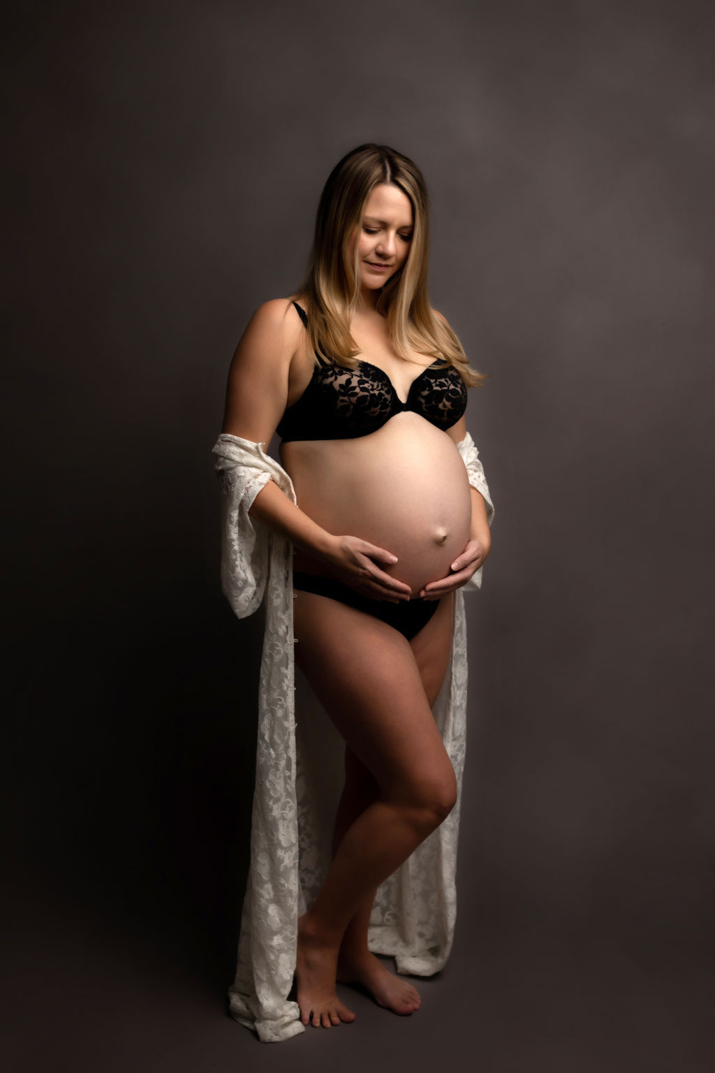 A mother to be in black underwear and a white coverup smiles down at her bump while standing in a studio
