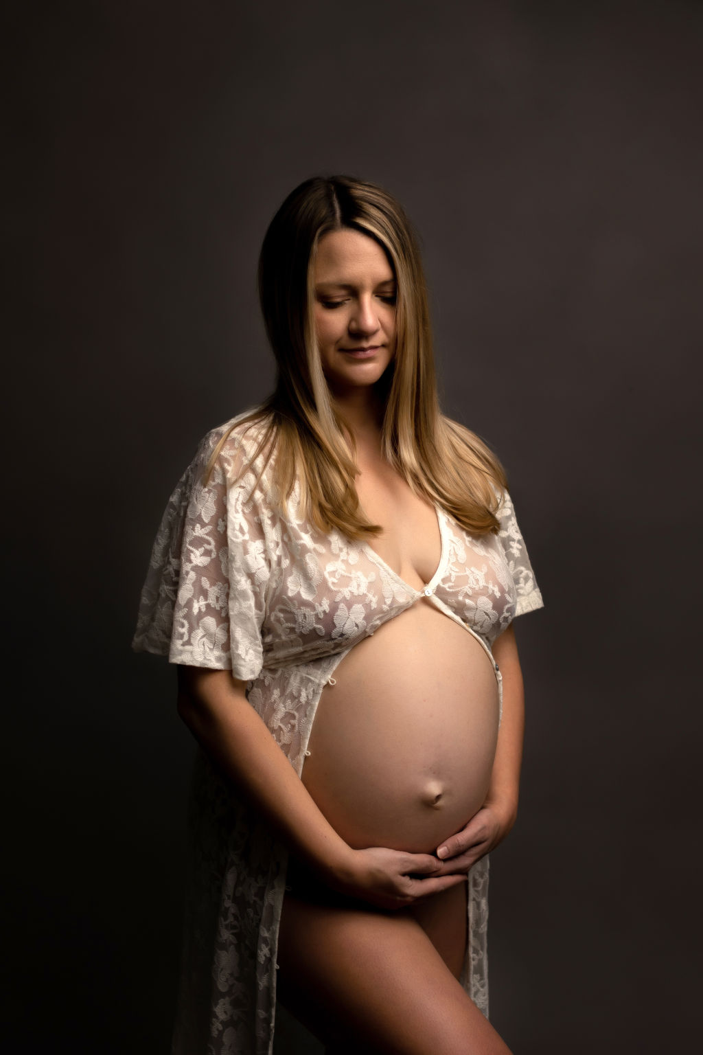 A mother to be stands in a studio in a white lace coverup holding her bump after a prenatal massage whitby