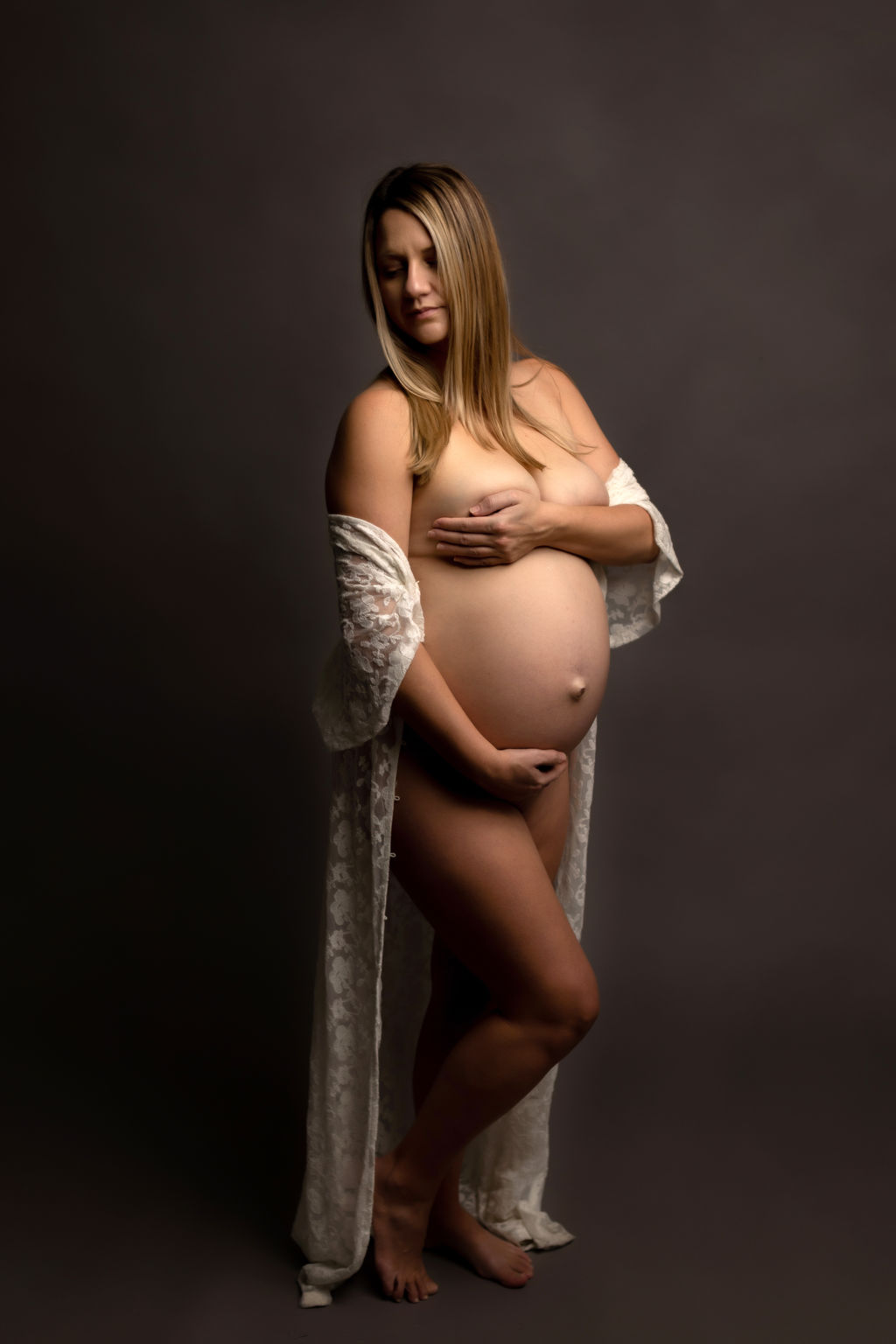 A mother to be stands in a studio wearing an open lace coverup and covering with her hands on bump and chest after a prenatal massage whitby