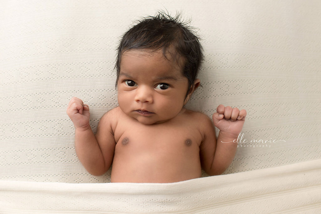 A newborn baby lays on a bed with eyes open and arms up